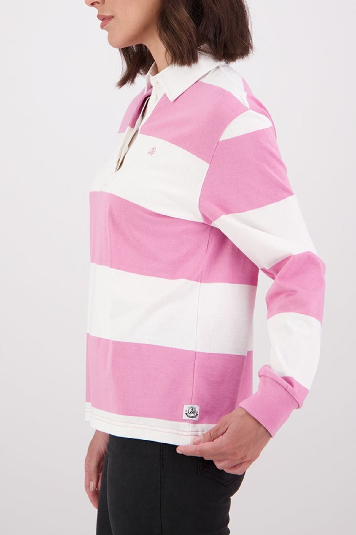 A woman in profile wears the Swanndri Ridgeway Womens Rugby, a 100% cotton shirt with wide pink and white stripes and a classic white collar, paired with black pants, against a plain light background.