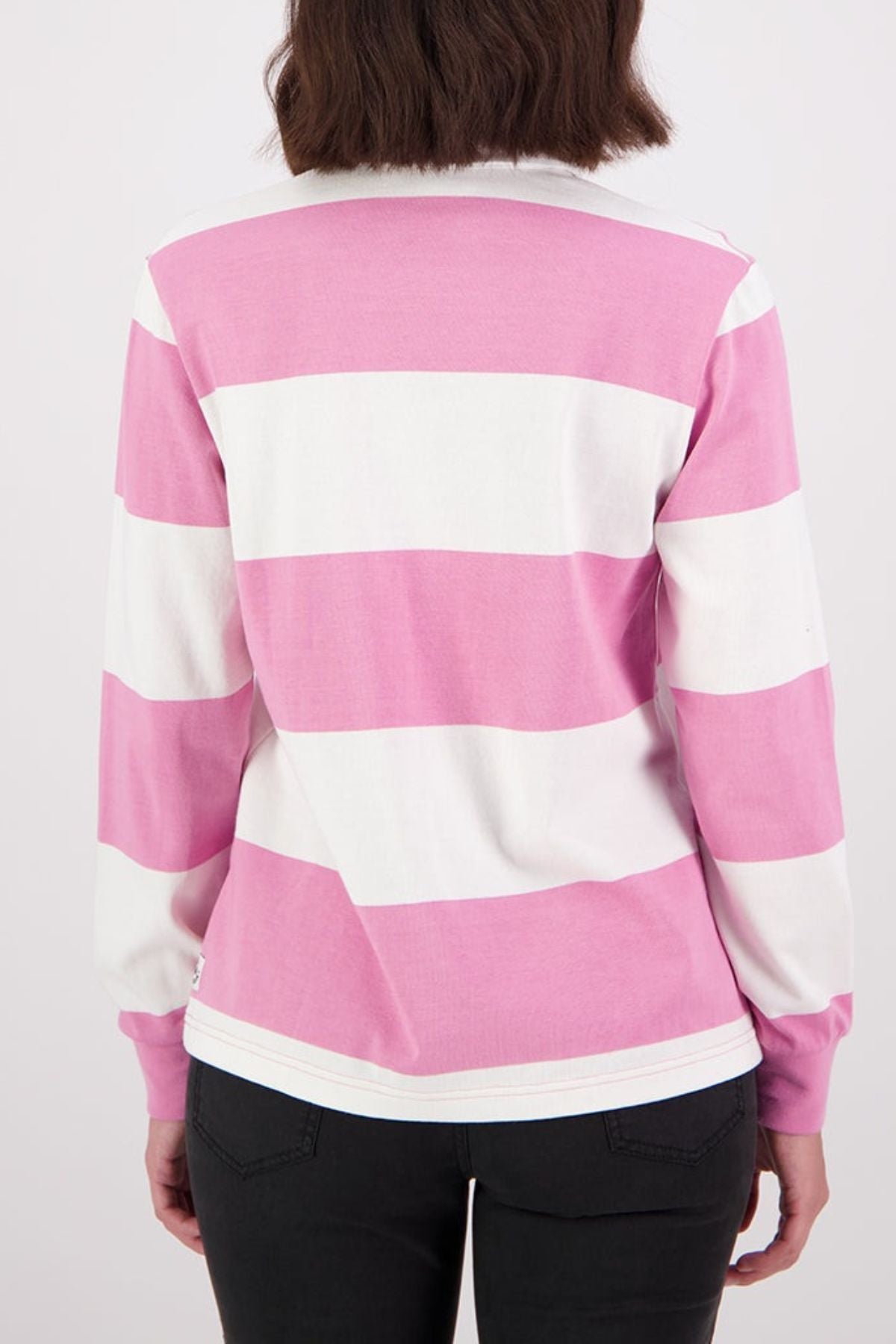 A person with shoulder-length brown hair is seen from behind wearing the Swanndri Ridgeway Womens Rugby, a 100% cotton long-sleeve shirt with wide pink and white stripes, paired with black pants.