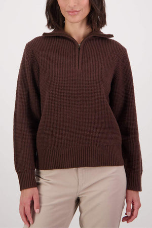 A person wears the Swanndri Riverside v2 Zip Neck Rib Jumper in brown, featuring a half-zip collar and textured knit, paired with light beige pants, while standing against a plain white background.