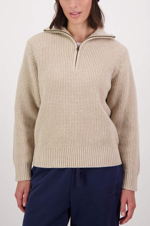 A woman wears a beige Swanndri Riverside v2 Zip Neck Rib Jumper with a collar, paired with dark blue pants, standing against a plain light background.