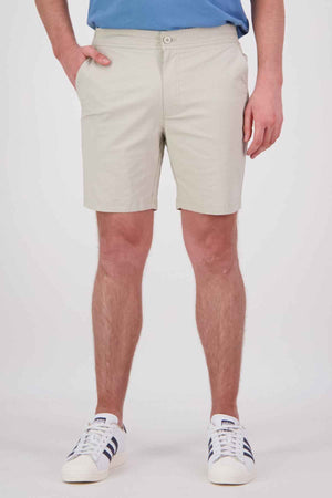 A person wearing Swanndri Sandy Point Shorts in a light beige color made from stretch cotton fabric, paired with a blue shirt and white sneakers featuring black stripes, stands with their hands in pockets against a plain background.