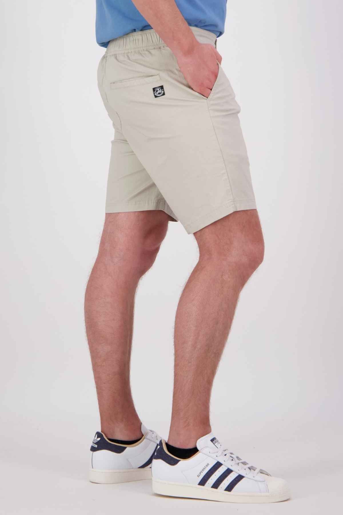 The individual in a blue shirt is wearing the Swanndri Sandy Point Shorts, featuring an elastic waistband and crafted from stretch cotton fabric in a beige tone. These shorts include a hidden zip back patch pocket. Matched with white sneakers accented by dark blue stripes, the person stands against a plain white background with their right hand slipped into one of the shorts' pockets.