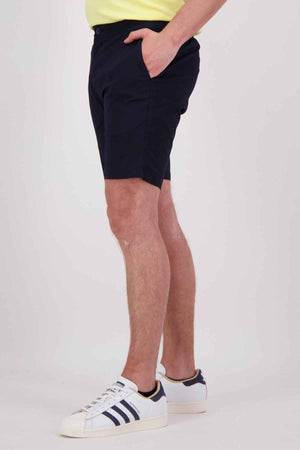 A person is standing against a plain background wearing Swanndri Sandy Point Shorts in navy blue, crafted from cotton stretch fabric. They have paired these shorts with a light-colored shirt and white sneakers that feature blue stripes. One of their hands rests in one of the multiple pockets of the shorts.