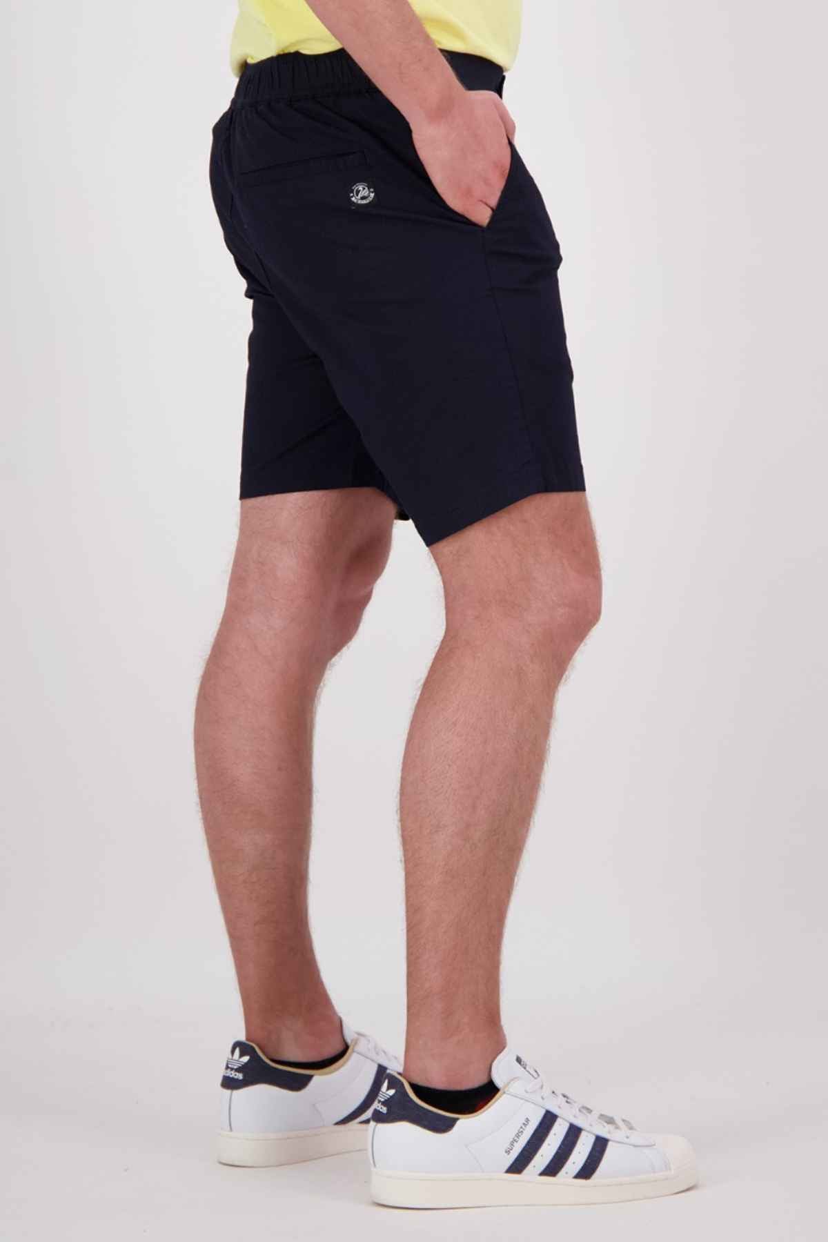 A person standing sideways is dressed in a yellow shirt, paired with the Swanndri Sandy Point Shorts, which feature multiple pockets. They wear white sneakers with blue stripes and casually rest their left hand in one of the shorts' pockets against a light-colored background.