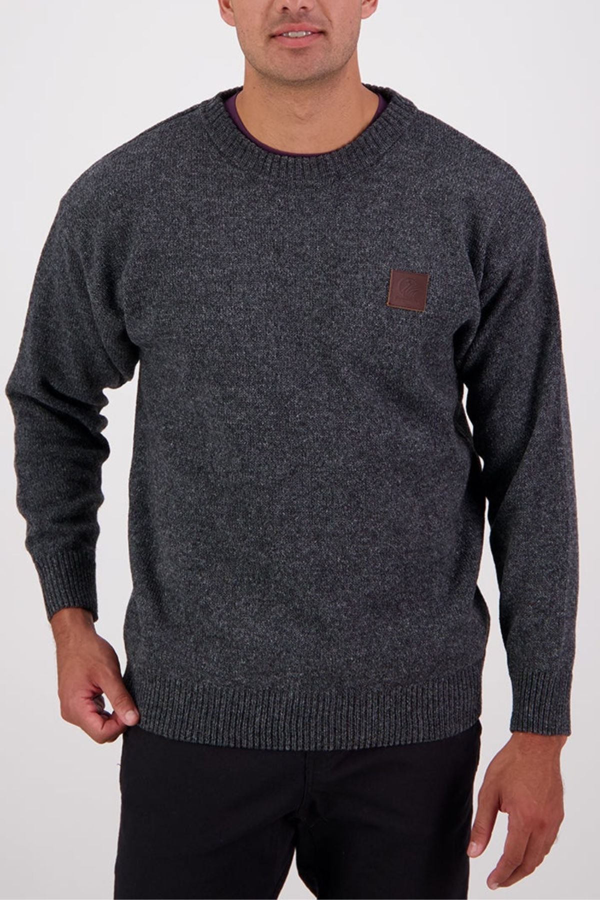 A man wears the Swanndri Seafarer Crew Neck Sweater—a dark gray, long-sleeve classic pullover with a small brown chest patch—paired with black pants, standing against a plain light background.