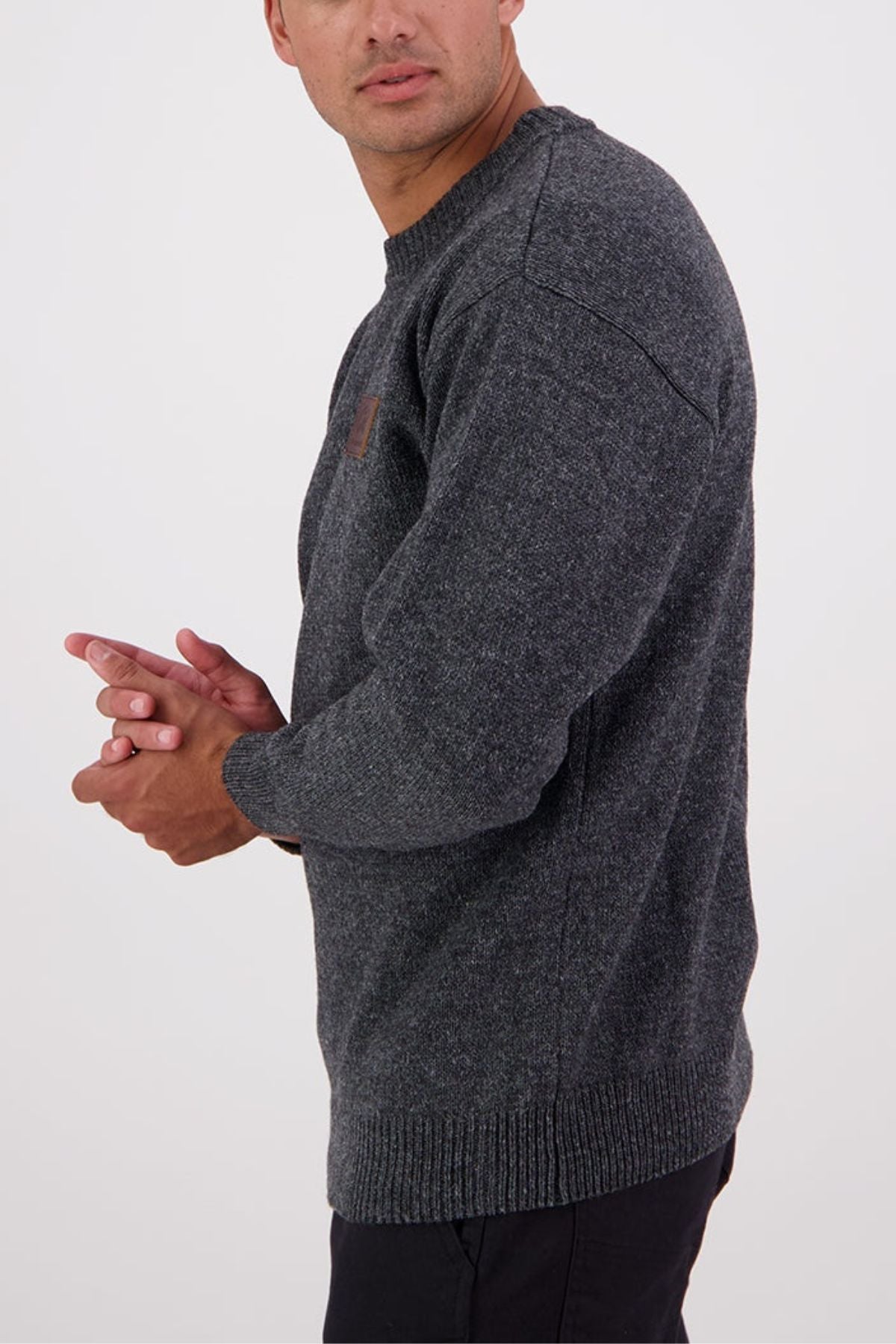 A man stands against a plain light background, angled to the side with hands together, wearing the Swanndri Seafarer Crew Neck Sweater and black pants.