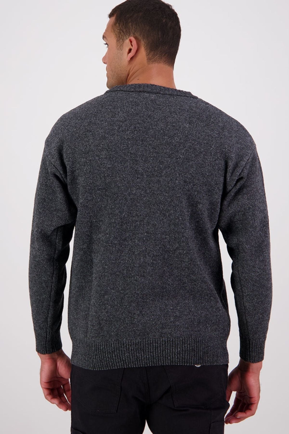 A man faces away from the camera, wearing the Swanndri Seafarer Crew Neck Sweater—a dark gray New Zealand wool sweater—with black pants, set against a plain, light background.