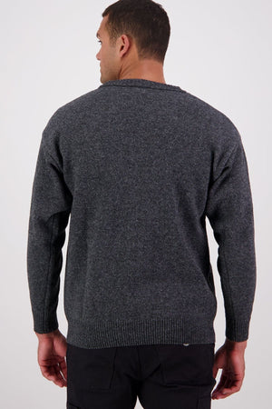 A man faces away from the camera, wearing the Swanndri Seafarer Crew Neck Sweater—a dark gray New Zealand wool sweater—with black pants, set against a plain, light background.