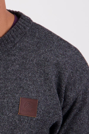 Close-up of a person wearing the Swanndri Seafarer Crew Neck Sweater in dark gray New Zealand wool, featuring a ribbed crew neck and a brown leather patch with the Swanndri logo on the chest. Only the upper torso is shown.