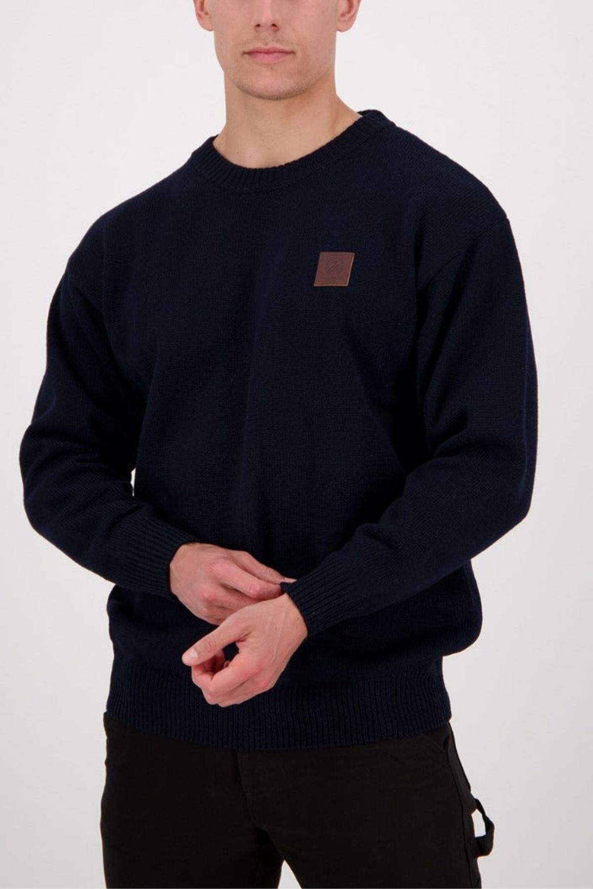 A person stands against a plain white background, hands clasped, wearing the Swanndri Seafarer Crew Neck Sweater in dark navy New Zealand wool with a small brown chest patch. This classic pullover boasts a timeless crew neck design.