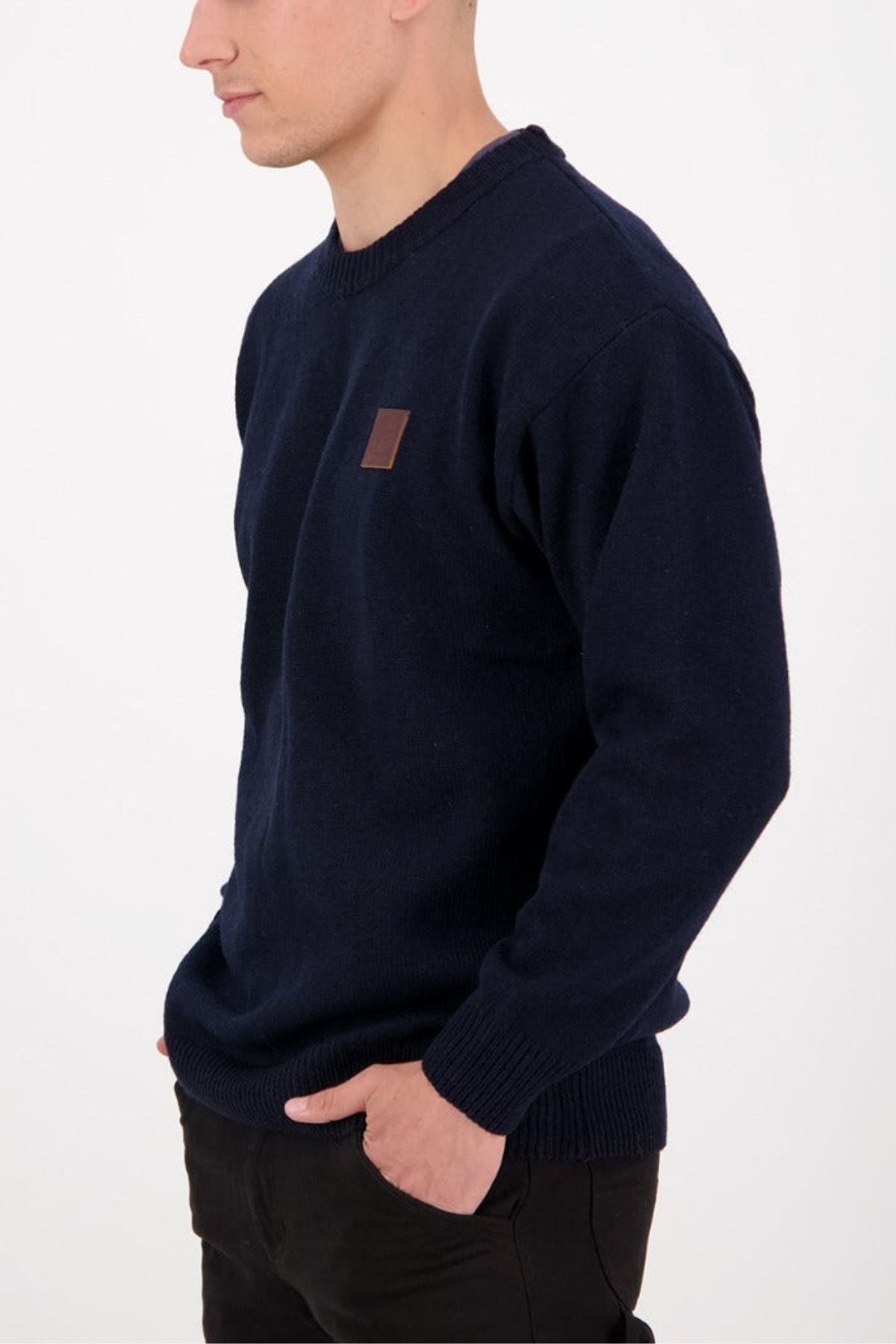 A person wears the Swanndri Seafarer Crew Neck Sweater in dark blue, featuring a small square brown chest patch, standing against a plain white background with one hand in their pocket.