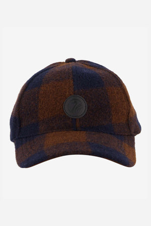 Check out the Swanndri Swanni Wool Check Baseball Cap with its dark brown and navy checkered pattern, rounded crown, curved brim, and circular black logo patch. It offers an adjustable buckle for the perfect fit against a plain white background.