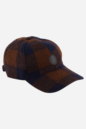 Introducing the Swanndri Swanni Wool Check Baseball Cap, showcasing a brown and dark blue checkered pattern with a stylish curved brim and a black circular logo on the front. It includes an adjustable buckle at the back, ensuring comfort and timeless style.