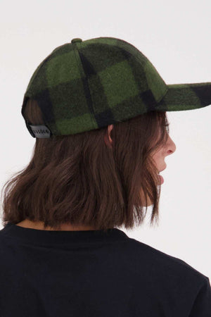 A person with shoulder-length brown hair wears a black shirt and a green and black Swanndri Swanni Wool Check Baseball Cap, shown from the back and side view, against a light background.