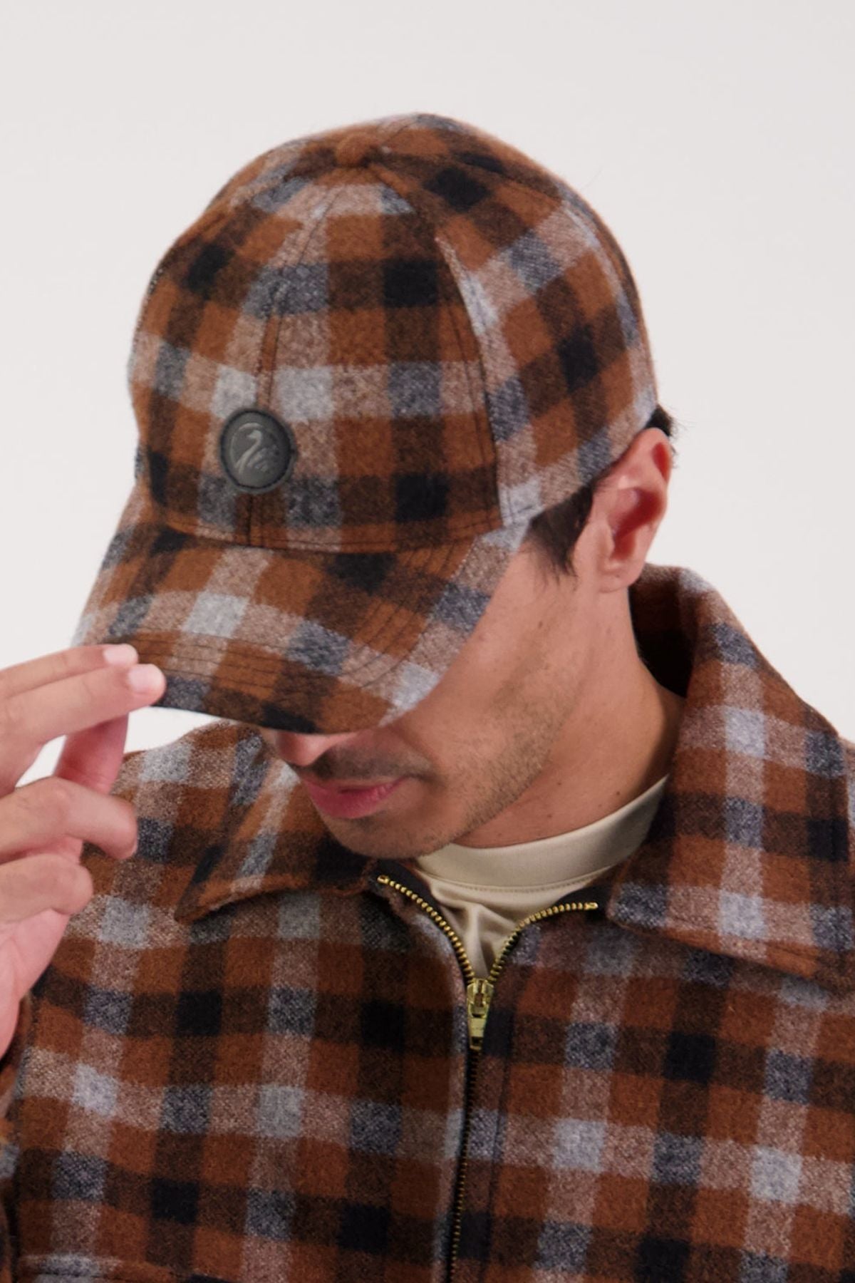 A person in a Swanndri brown, black, and gray plaid jacket and the Swanndri Swanni Wool Check Baseball Cap holds the cap's brim, looking down to partly hide their face against a plain light backdrop.