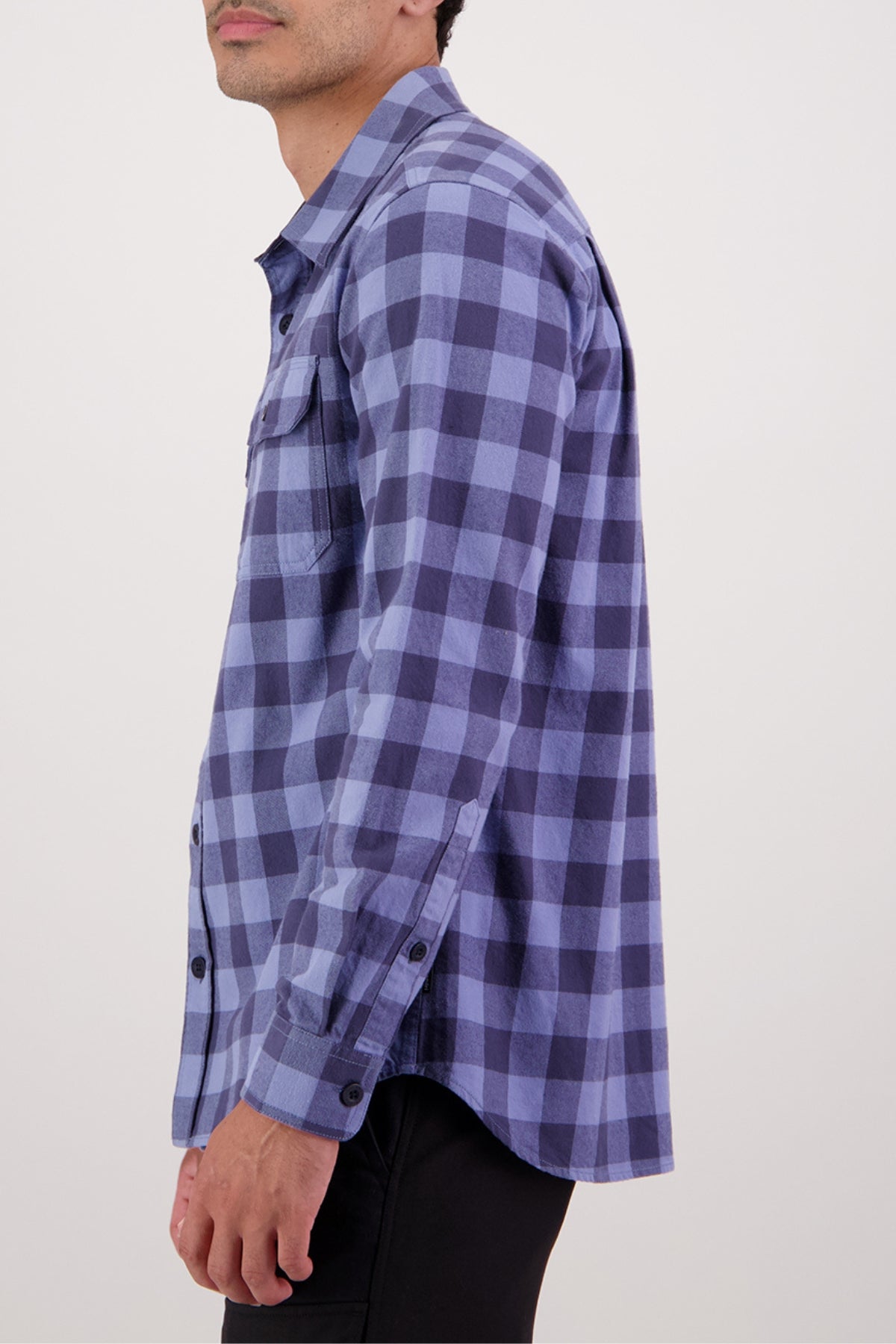 A person is seen from the side against a plain background wearing the Swanndri Taranaki Tailor Shirt in blue and navy plaid, a 100% cotton style with black buttons and buttoned chest pockets.