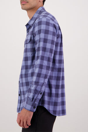 A person is seen from the side against a plain background wearing the Swanndri Taranaki Tailor Shirt in blue and navy plaid, a 100% cotton style with black buttons and buttoned chest pockets.