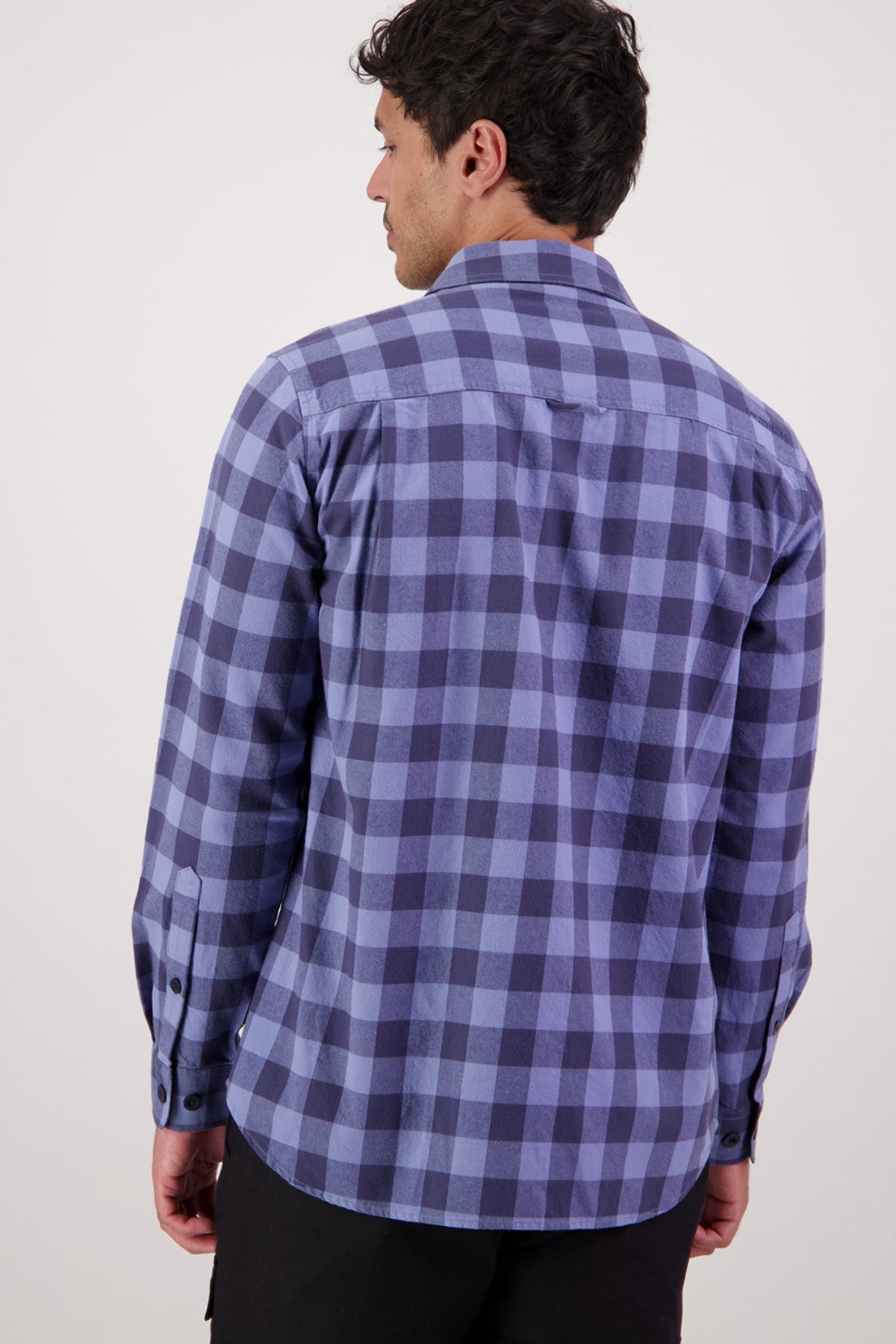 A man is seen from the back wearing a Swanndri Taranaki Tailor Shirt—long-sleeved, blue and navy checkered, 100% cotton with buttoned chest pockets—and black pants, set against a plain light background.