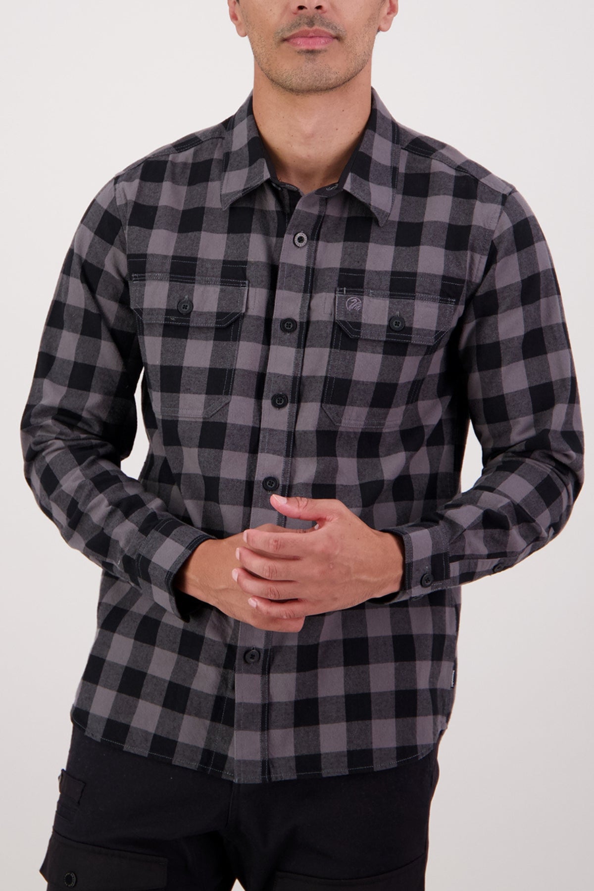 A man stands against a plain background wearing the Swanndri Taranaki Tailor Shirt, a men's long-sleeve black and gray plaid shirt made of 100% cotton, showcasing its stylish and comfortable design with his hands clasped in front.