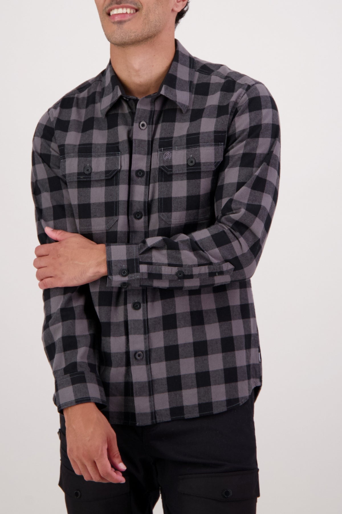 A person in a Swanndri Taranaki Tailor Shirt—a long-sleeve, black and gray men's casual shirt—stands against a plain light background with one arm crossed, smiling slightly.