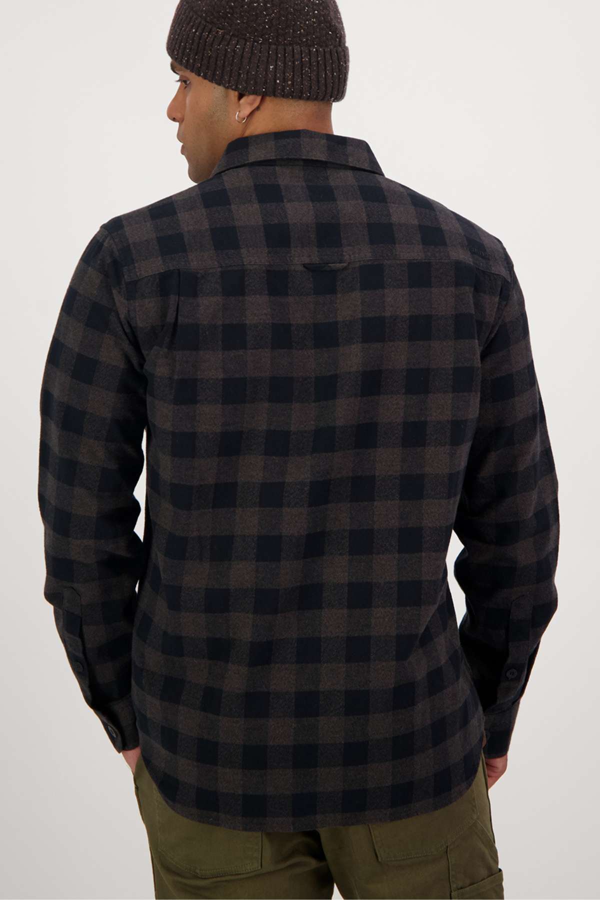 A person dressed in a Swanndri Taranaki Tailor Shirt featuring a dark checkered pattern, green pants, and a brown knit beanie is seen from behind. With their hands resting at their sides, they stand against a plain white background.