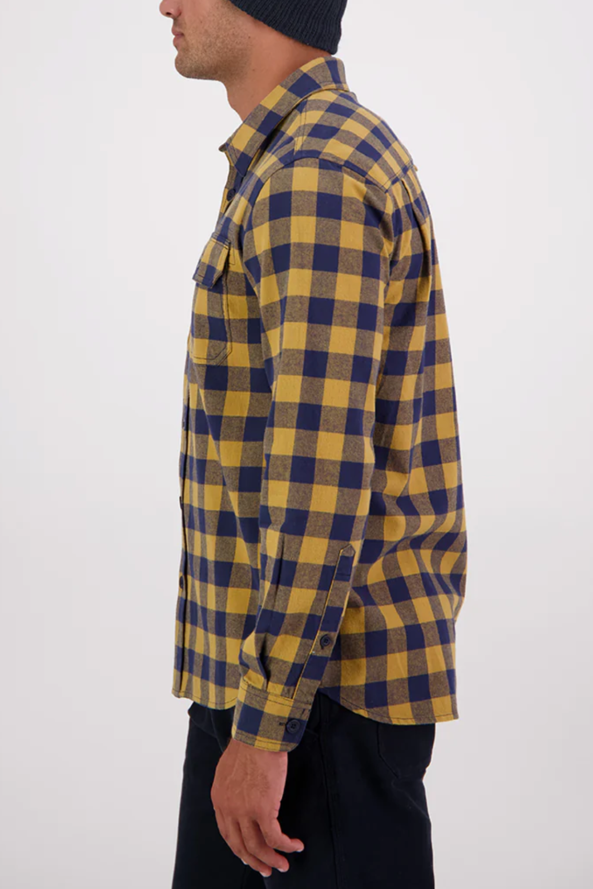 Someone wearing a yellow and navy blue plaid Swanndri Taranaki Tailor Shirt with buttoned chest pockets and dark pants stands sideways, facing left, against a plain white background.