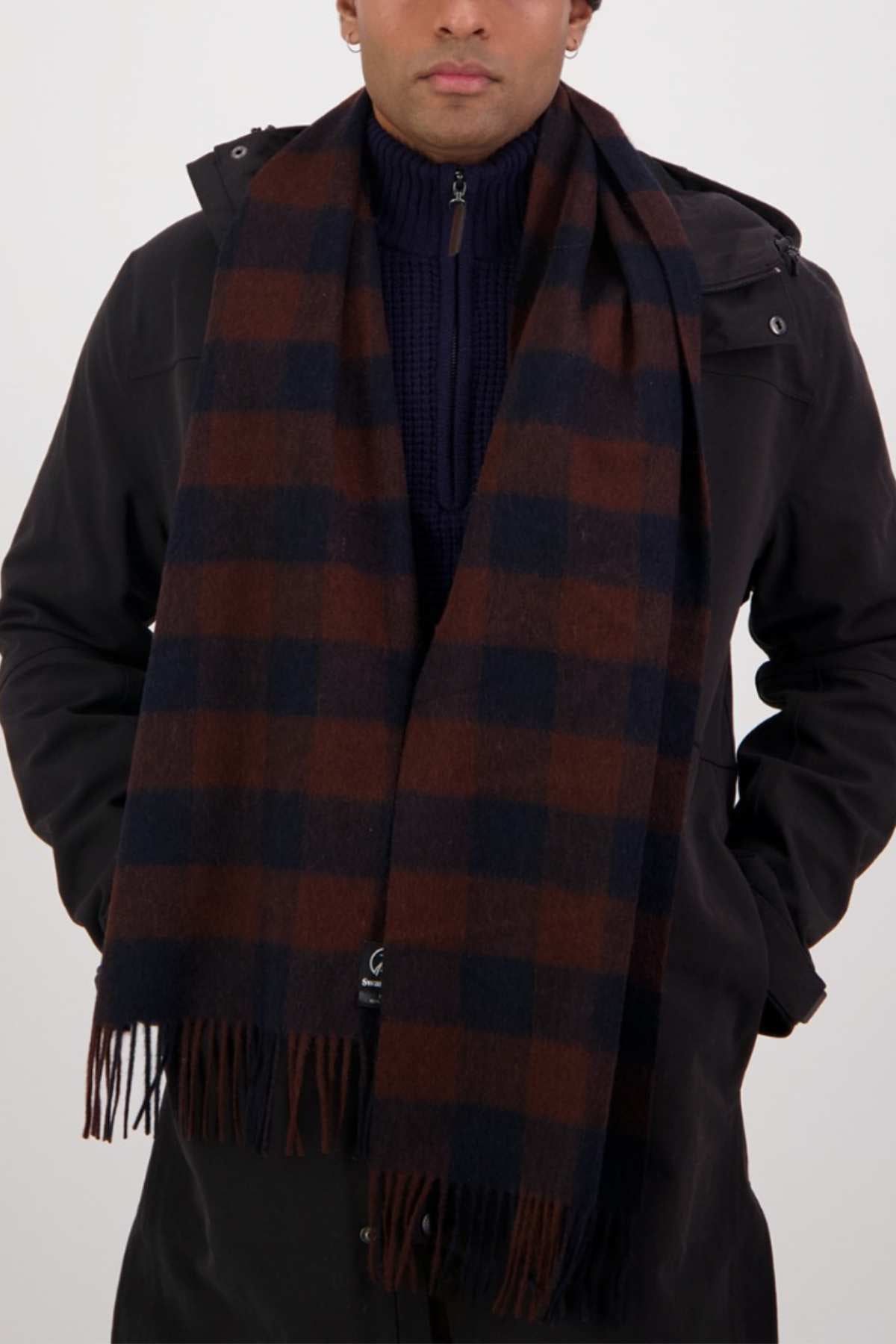 A person in a dark coat and navy blue sweater wears the Swanndri The Scarfie—a brown and navy checkered brushed wool scarf with fringe—around their neck. The cold weather accessory partially obscures their face.