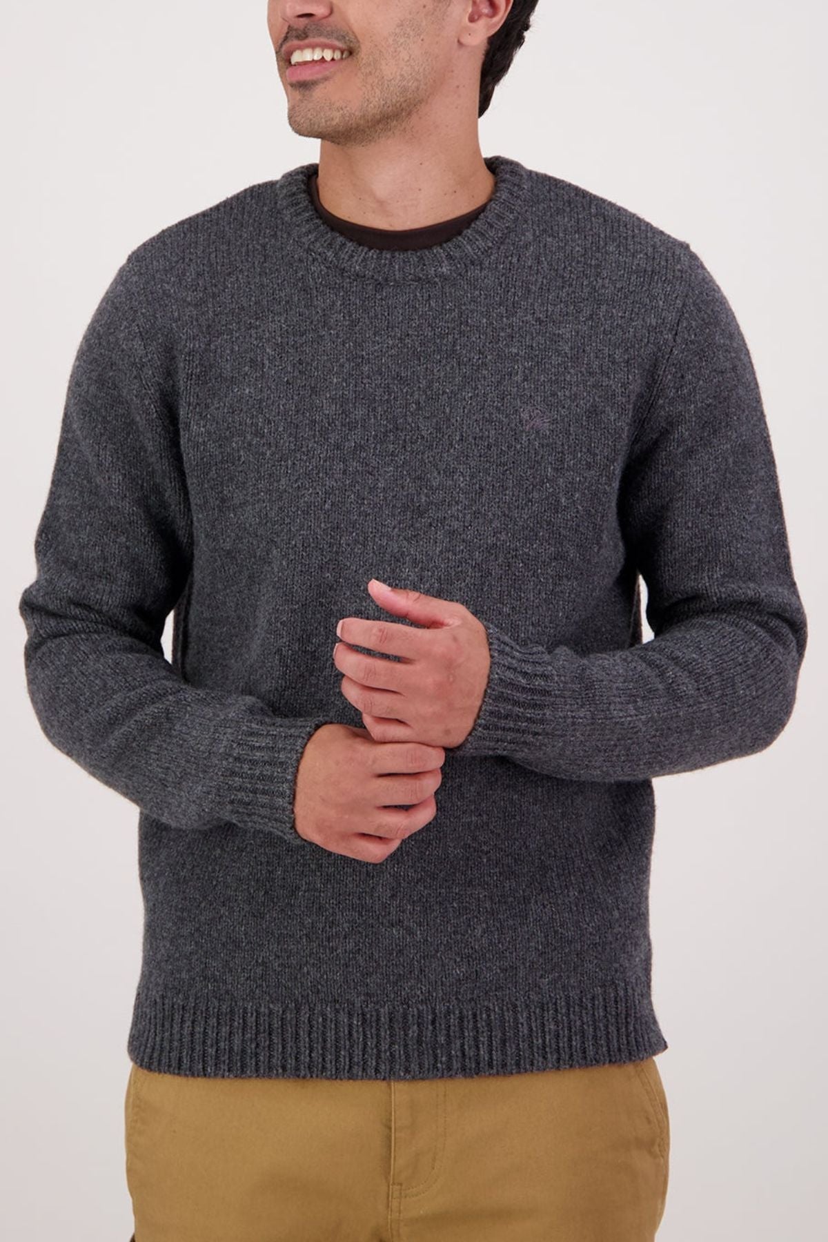 Wearing the Swaandri Waylins Wool Knit Crew in gray over a dark shirt and tan pants, a person stands against a plain light background, looking to the side with hands clasped in front.