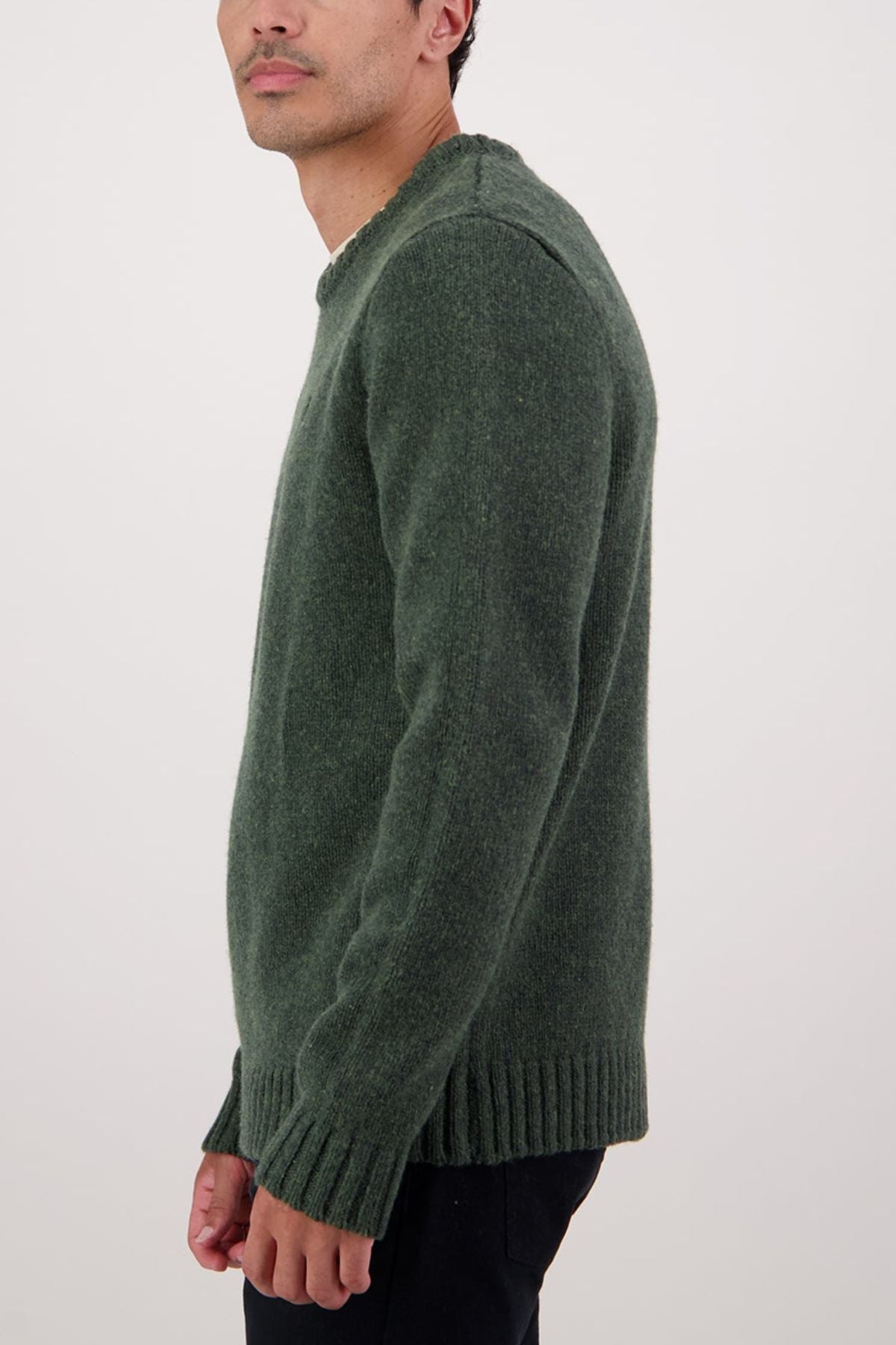 A person stands against a plain light background, wearing a dark green Swaandri Waylins Wool Knit Crew and black pants, shown from the side with their head slightly turned and only part of their face visible.