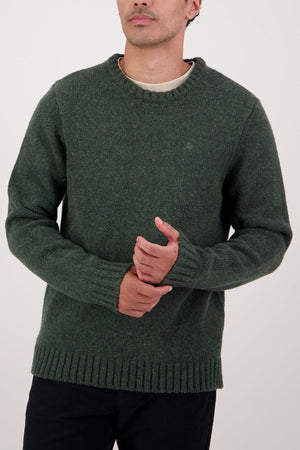 A person in a dark green Swaandri Waylins Wool Knit Crew and black pants stands against a plain light background, hands clasped. The durable crew neck knitwear is visible, with their face partially out of frame.