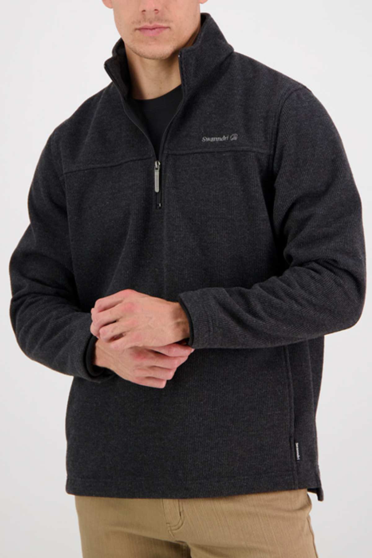 Man wearing a Swanndri Weka Sweater, a heavyweight dark gray zip-up made from breathable windproof material, over a black shirt and beige pants. It has a small embroidered chest logo against a plain white background with his hands clasped in front.