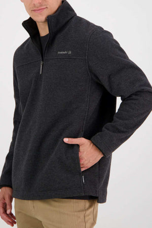 Wearing a dark gray Swanndri Weka Sweater—breathable, windproof quarter-zip fleece—with one hand in the pocket and light brown pants, the person stands against a plain light background.