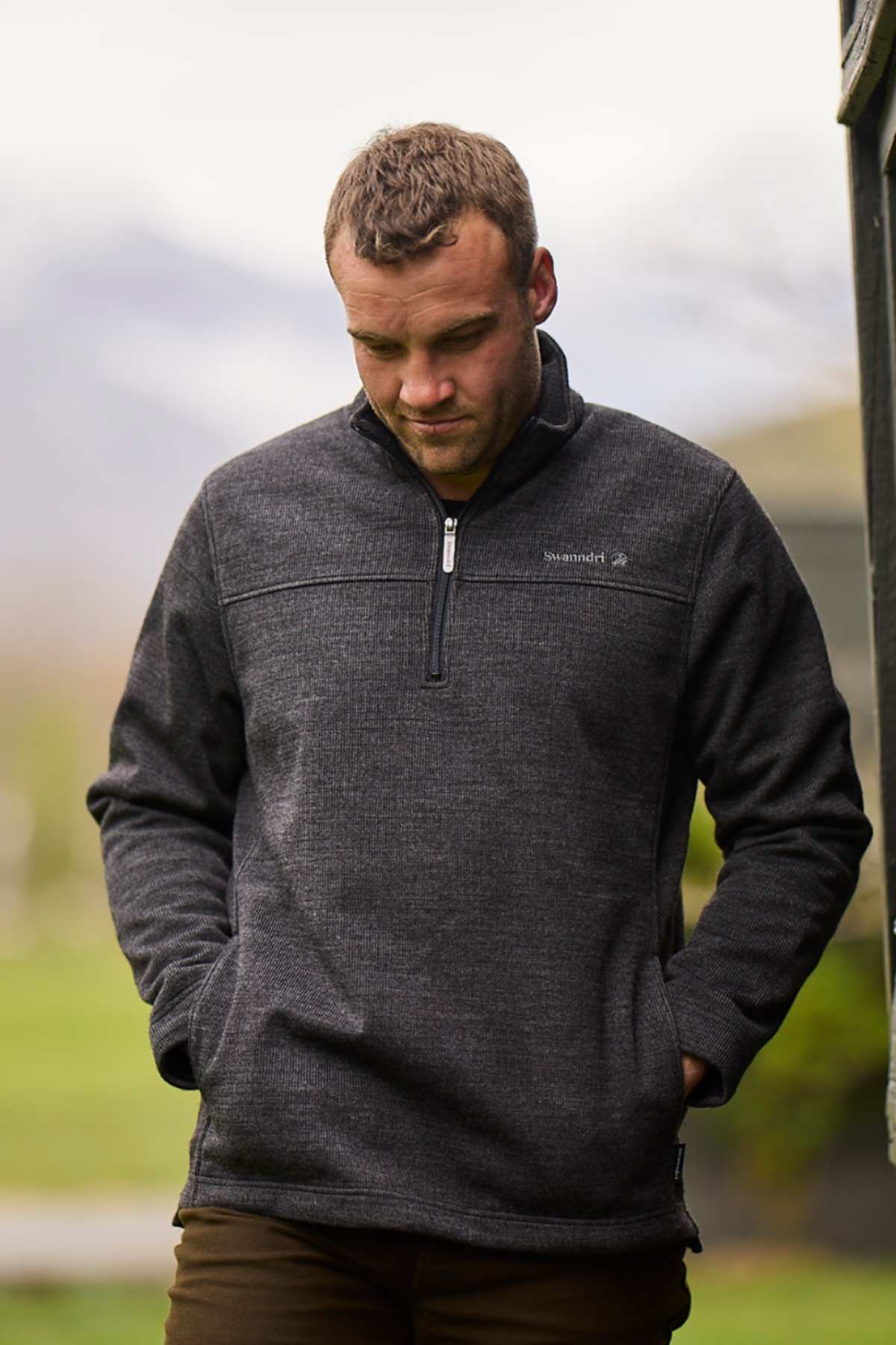 A man with short hair stands outdoors, hands in his pockets, wearing the dark gray Swanndri Weka Sweater. He looks down against a softly blurred green background that suggests a natural setting.
