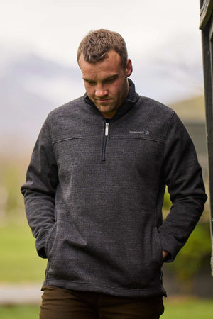 A man with short hair stands outdoors, hands in his pockets, wearing the dark gray Swanndri Weka Sweater. He looks down against a softly blurred green background that suggests a natural setting.