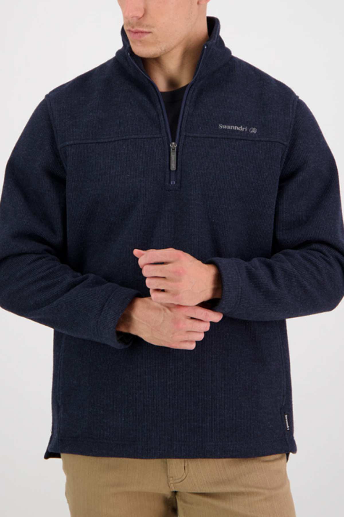 A person wearing a Swanndri Weka Sweater in navy blue with a small logo on the left chest and tan pants. Their face is partially turned to the side, and they have their hands lightly clasped in front of them. The background is plain white.