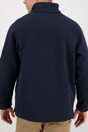 A person is seen from the back wearing a Swanndri Weka Sweater, a dark navy blue, breathable and windproof jacket with a high collar. The sweater features visible seam lines running across the shoulders and down the sleeves. Complementing the outfit, they are also wearing light brown pants. The background is plain and light-colored.