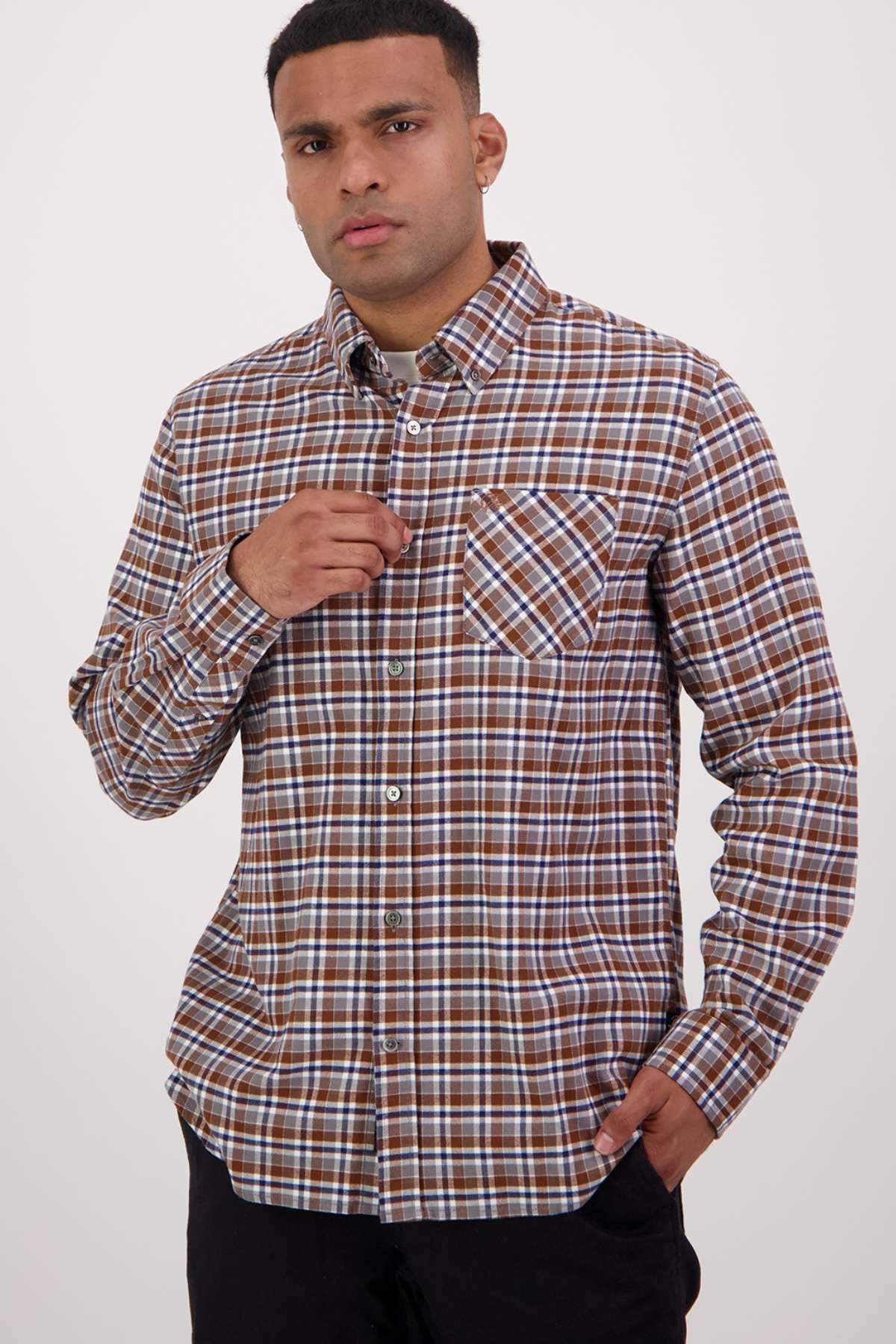A man stands against a plain background, wearing the Swanndri Wendon Shirt in classic fit, featuring a brown and white plaid pattern. The 100% cotton shirt sports long sleeves. His left hand is touching a button near his chest, while his right hand is tucked into his pocket. He has short hair and maintains a neutral expression.