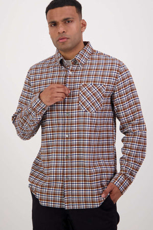 A man stands against a plain background, wearing the Swanndri Wendon Shirt in classic fit, featuring a brown and white plaid pattern. The 100% cotton shirt sports long sleeves. His left hand is touching a button near his chest, while his right hand is tucked into his pocket. He has short hair and maintains a neutral expression.