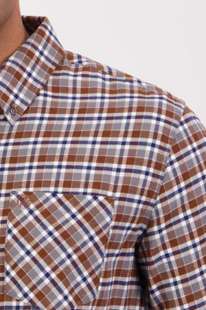Close-up of a person wearing a Swanndri Wendon Shirt in a brown, blue, and white plaid pattern. The shot captures only the upper torso and part of the neck, highlighting the intricate pattern and texture of the 100% cotton fabric. This classic fit shirt is detailed with a front pocket.