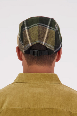 A person seen from behind wears a Swanndri Wentworth Valley v2 Wool Cap with an adjustable buckle and a light brown corduroy jacket. The background is plain and light-colored.