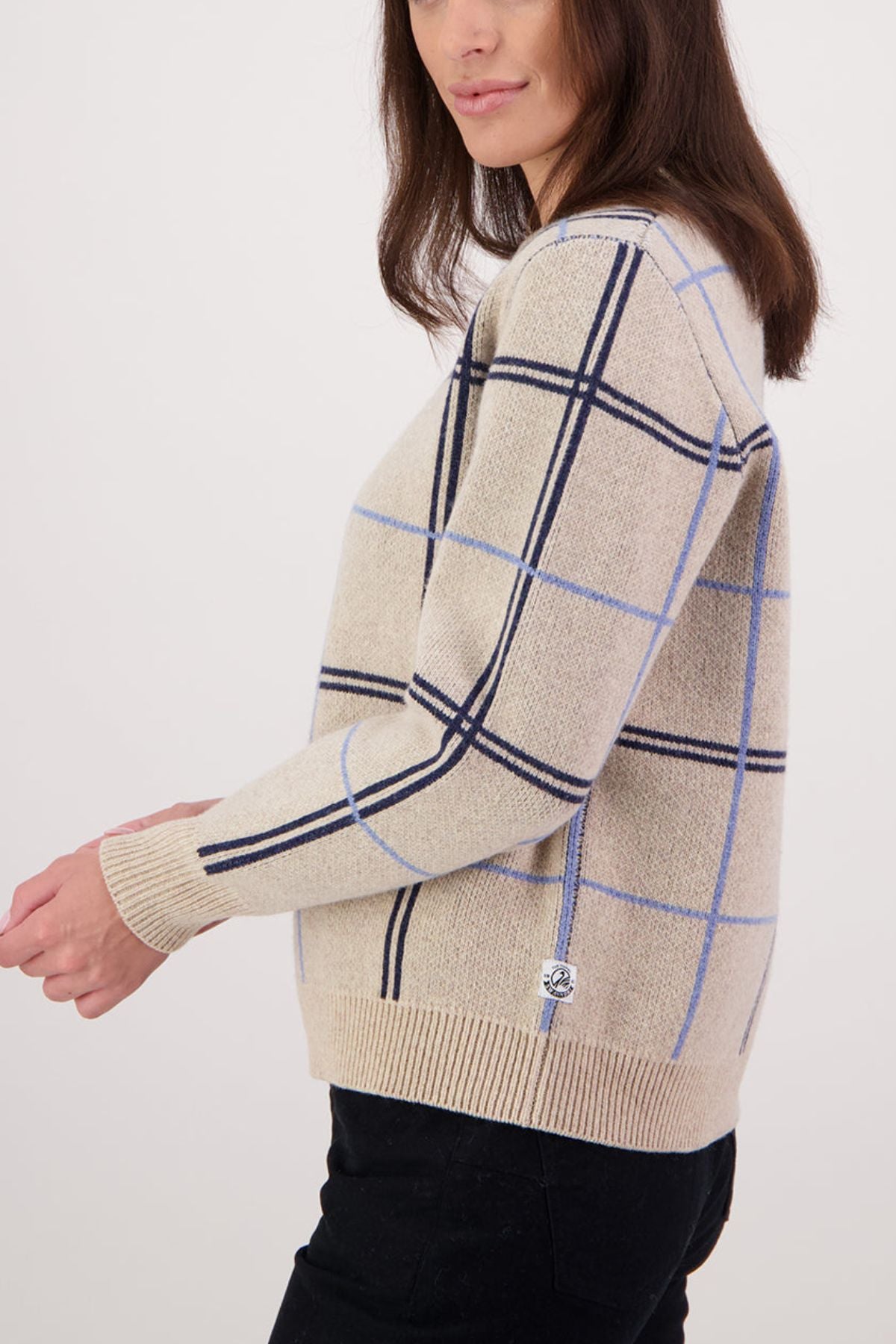 A person wears the Swanndri Yasmin Check Womens Knit, a beige merino wool sweater with black and blue plaid, paired with black pants, shown in profile against a plain light background.