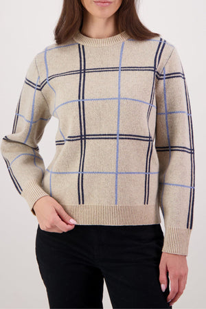 Wearing the Swanndri Yasmin Check Womens Knit in blue and black with black pants, a person stands against a plain light backdrop, their head partially out of frame.