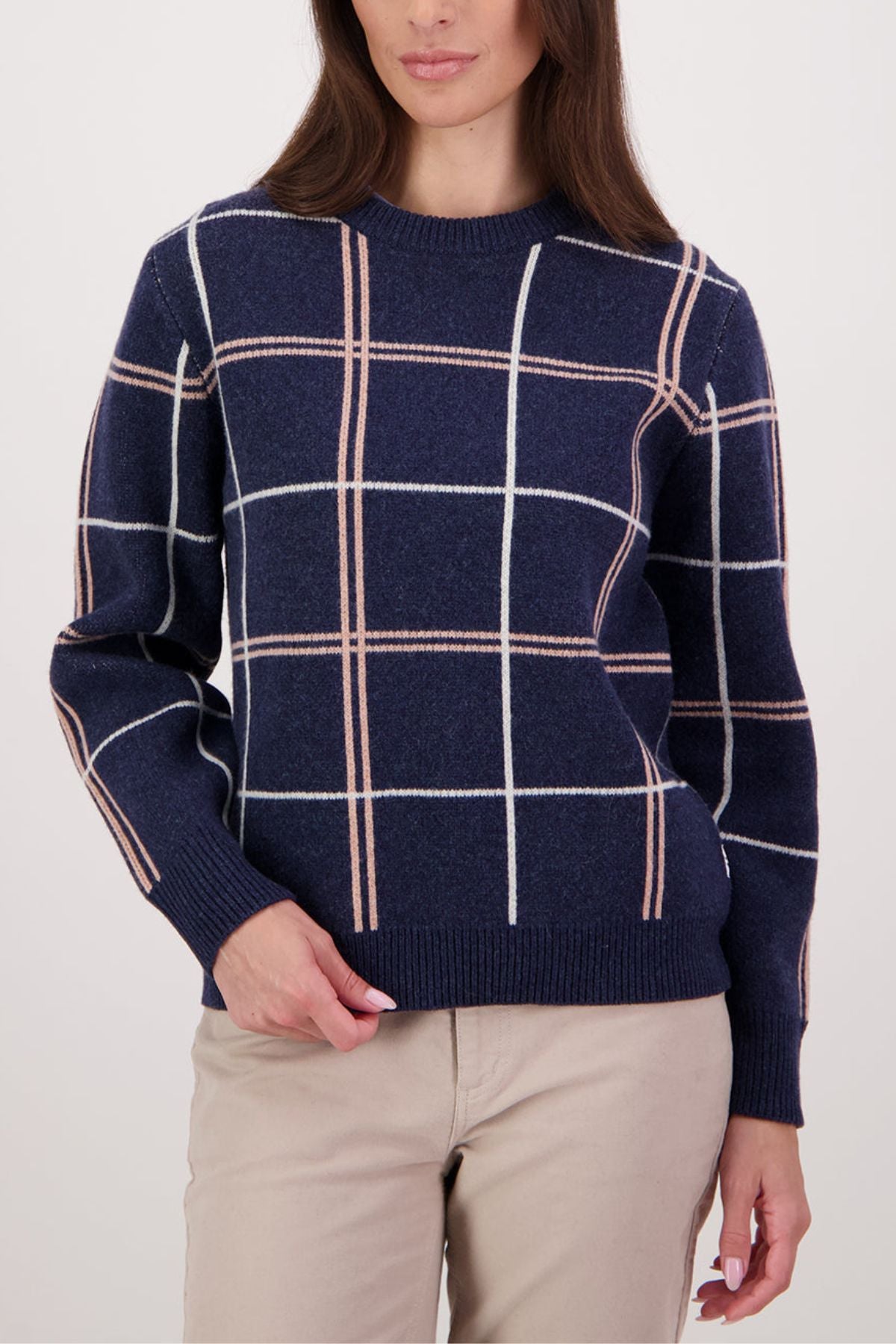 A woman stands against a light background wearing the Swanndri Yasmin Check Womens Knit in navy blue with white and light pink windowpane plaid, paired with beige pants.