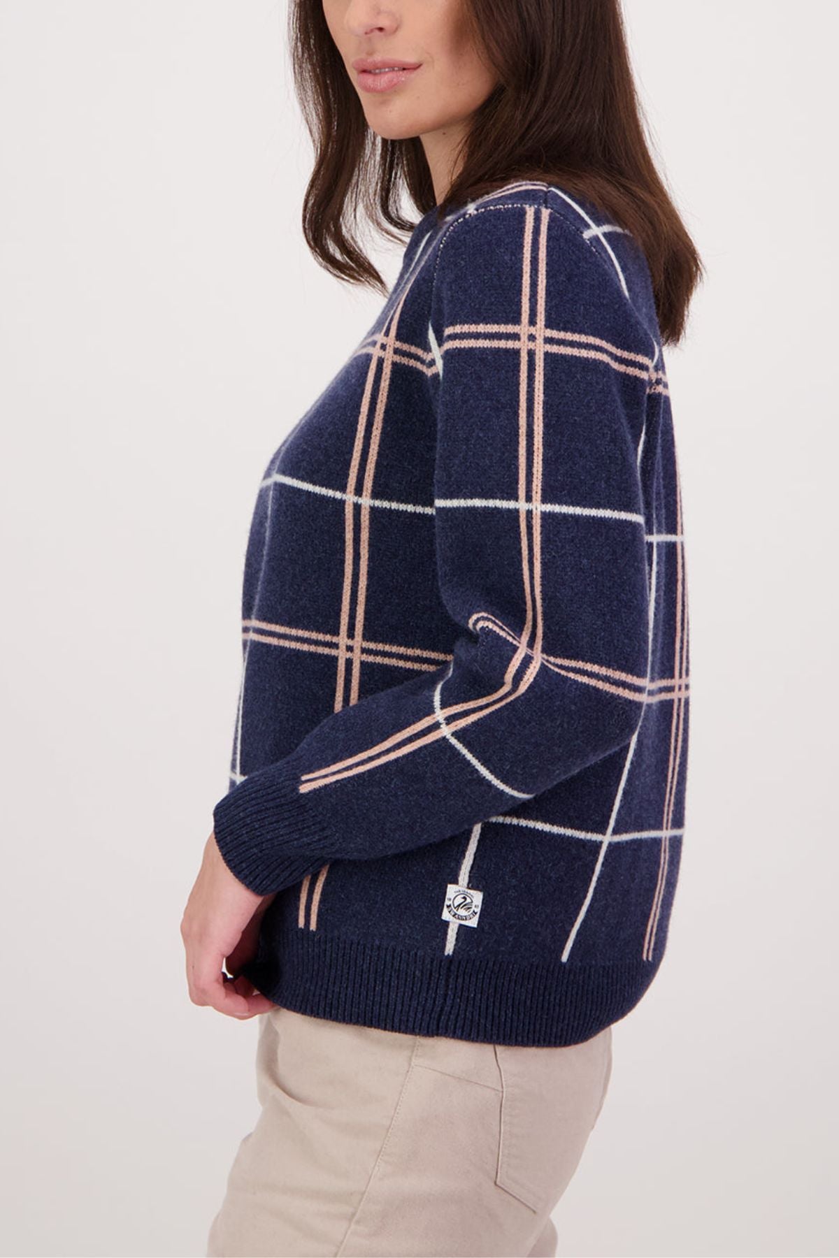 A woman in a Swanndri Yasmin Check Womens Knit navy blue merino sweater with white and beige windowpane pattern and light beige pants stands sideways against a plain white background.