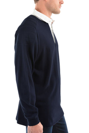 A man is shown from the side wearing a navy Thomas Cook Bill Rugby shirt with a white collared shirt underneath. The pullover features a short neck zipper and quilted shoulder panels for detail.