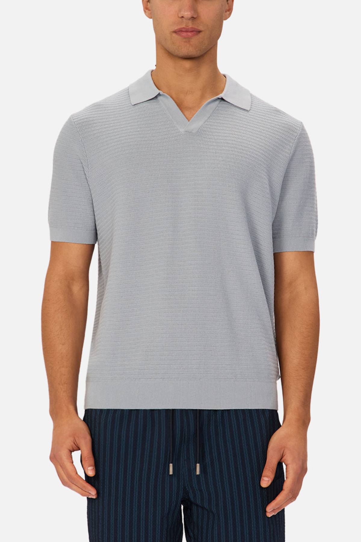 A man wearing the Industrie The Alicante Polo, a light gray knitted shirt with a small collar and v-neck, pairs it with dark blue pinstriped drawstring pants while standing against a plain white background.