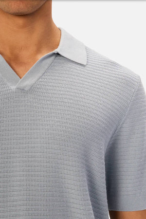 A person wearing the Industrie The Alicante Polo, a light blue men’s cotton polo with a textured knit pattern and open collar. Only their upper torso and part of their face are visible.