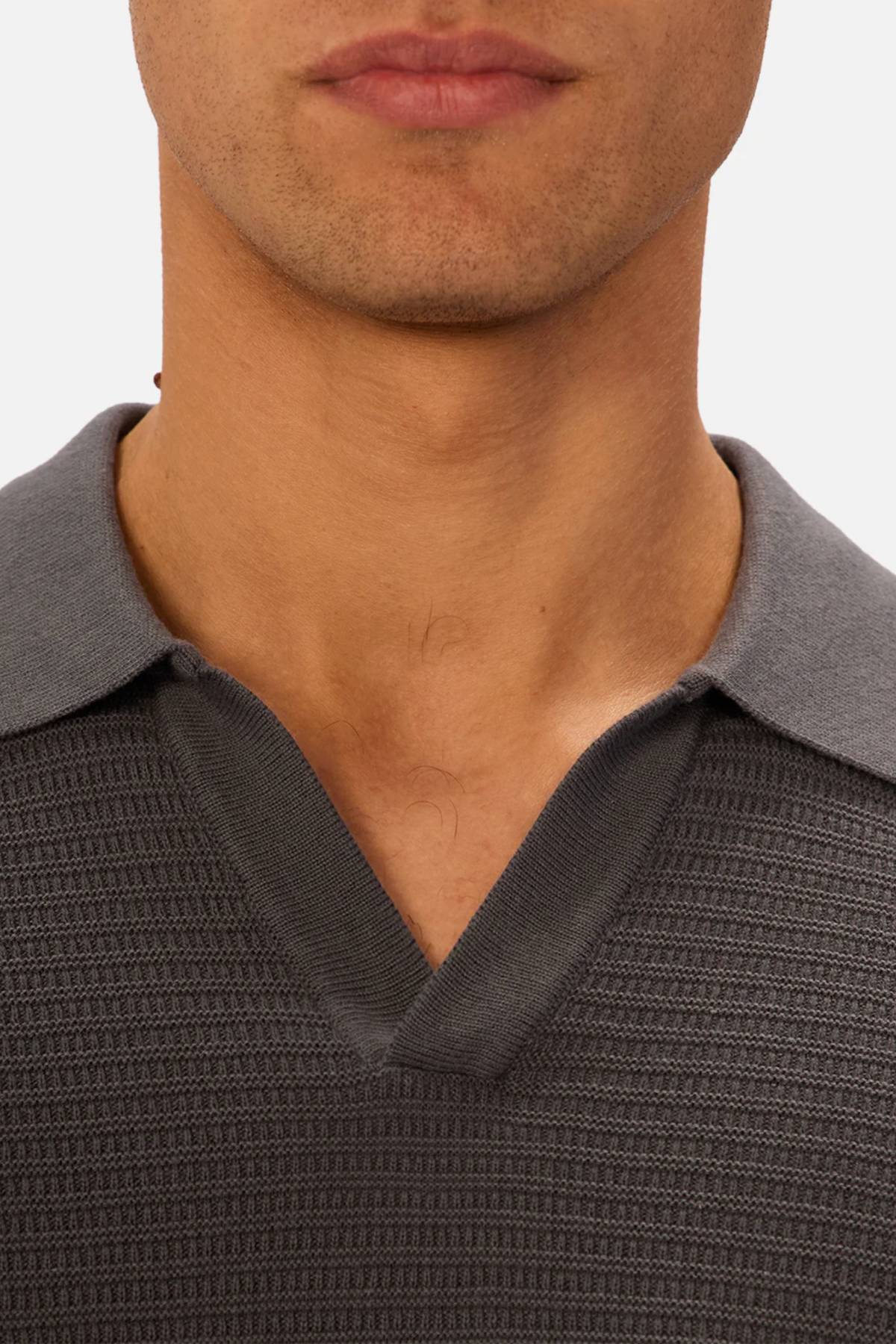 A close-up of a man's lower face and neck shows him wearing the Industrie The Alicante Polo, featuring a textured dark gray design with a light gray collar. The plain, light background highlights the polo's stylish, knitted look.