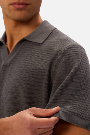 A man wears the Industrie The Alicante Polo, a textured, short-sleeve men's cotton shirt, gently holding his sleeve. The polo stands out against a plain, light-colored background.