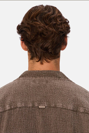 A person with short, wavy brown hair is shown from behind wearing the Industrie The Canyon Short Sleeve Shirt in textured brown, featuring a small metallic tag on the upper back. The background is plain and light-colored.